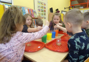 Dzieci nabierają pipetami zabarwione ciecze stojące w pojemnikach na środku stolika.